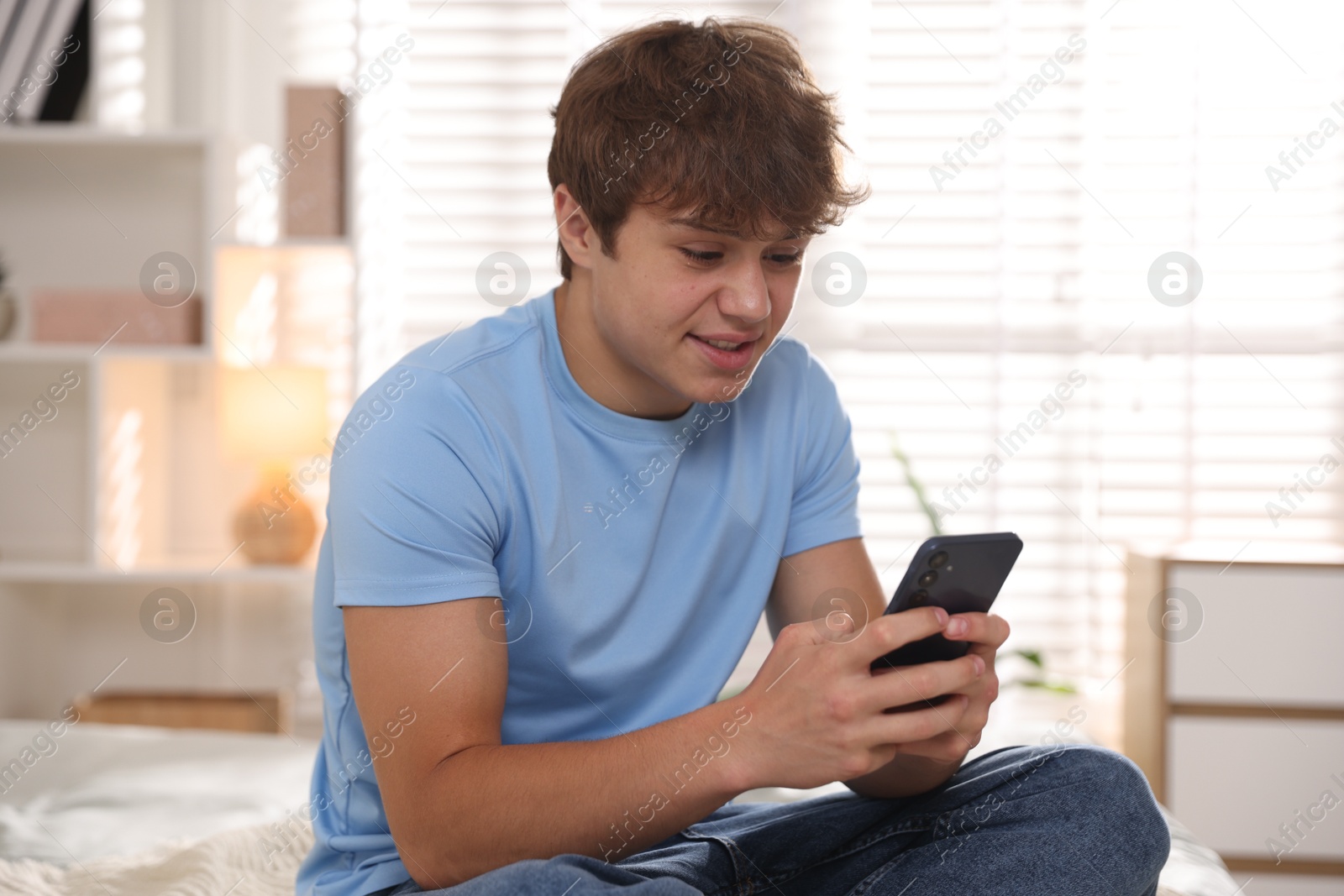 Photo of Smiling teenage boy using smartphone on bed at home