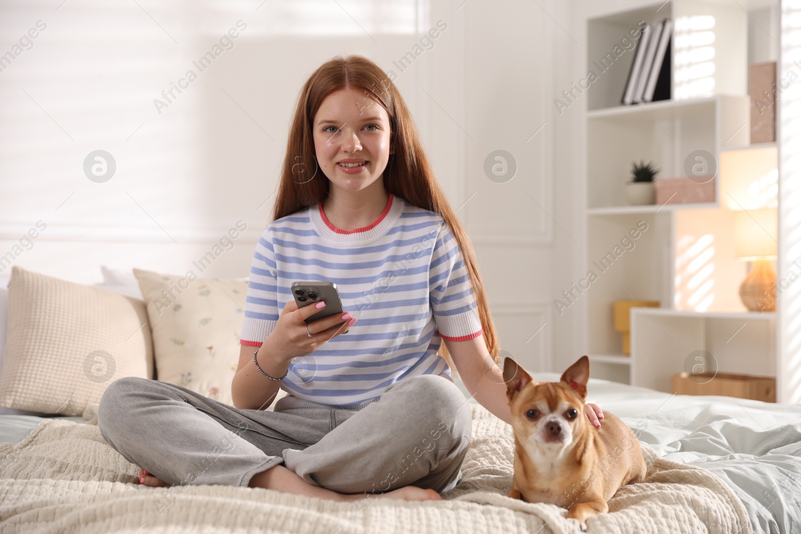 Photo of Smiling teenage girl using smartphone and dog on bed at home