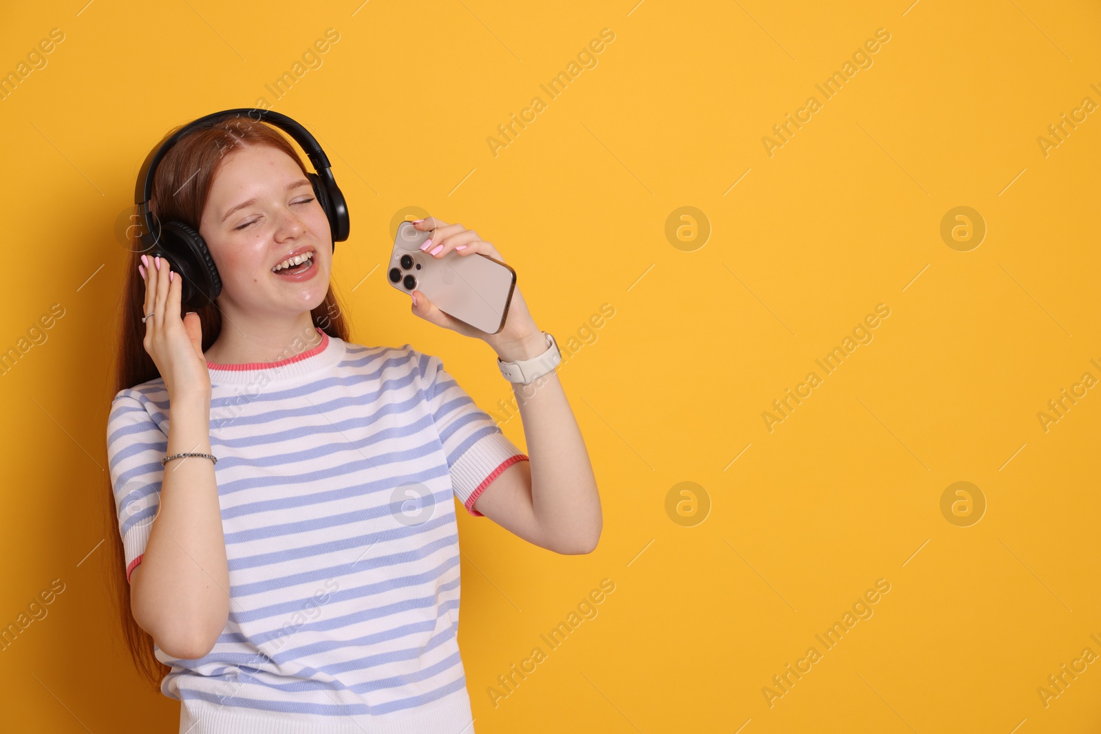 Happy teenage girl in headphones with smartphone singing on yellow background. Space for text Photo of Happy teenage girl in headphones with smartphone singing on yellow background. Space for text