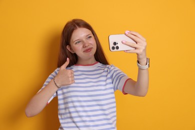 Photo of Beautiful teenage girl taking selfie by smartphone and showing thumbs up on yellow background
