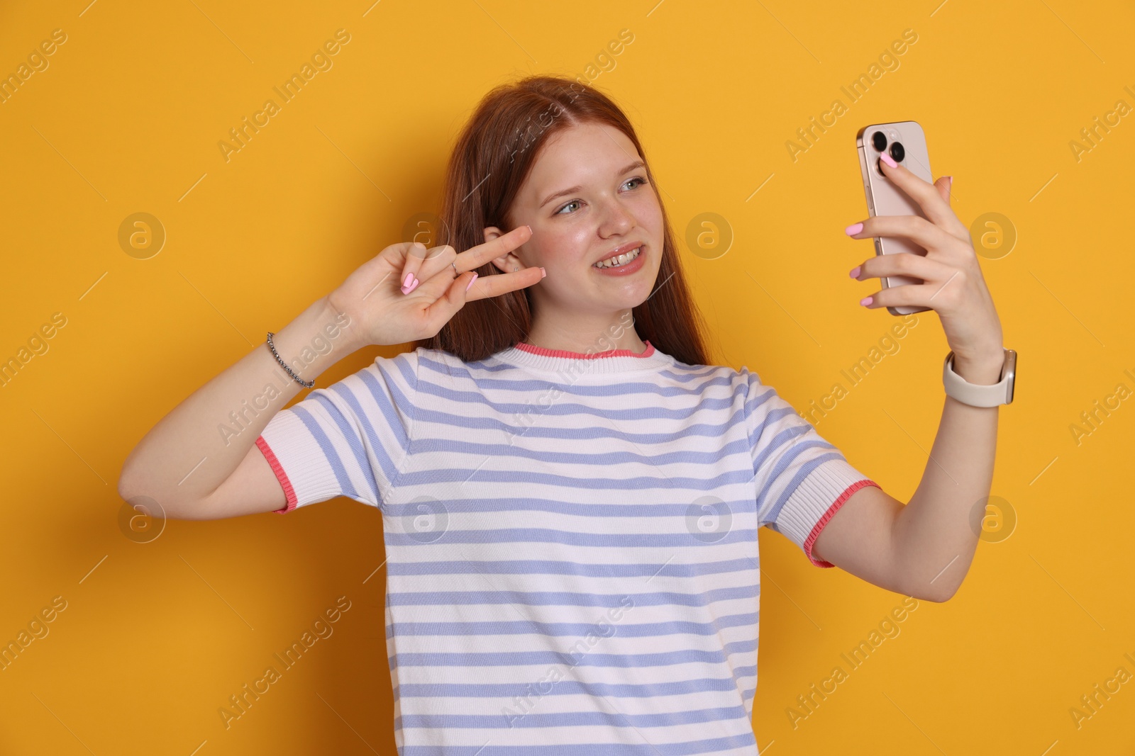 Photo of Smiling teenage girl taking selfie by smartphone and showing peace sign on yellow background