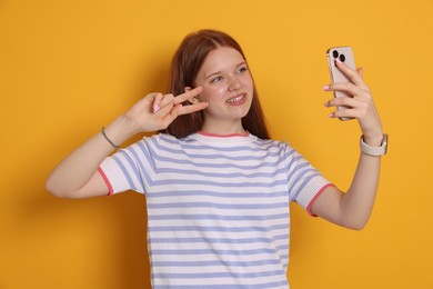 Photo of Smiling teenage girl taking selfie by smartphone and showing peace sign on yellow background