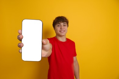 Photo of Smiling teenage boy showing smartphone with blank screen on yellow background, selective focus. Mockup for design