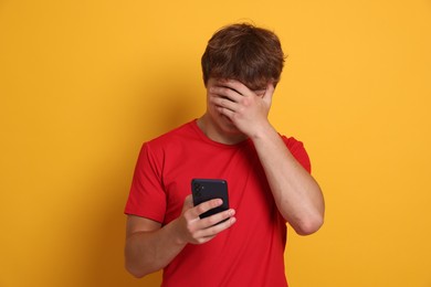 Photo of Teenage boy with smartphone covering face with hand on yellow background