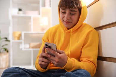 Photo of Teenage boy using smartphone near chest of drawers at home