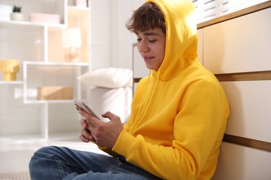 Photo of Teenage boy using smartphone near chest of drawers at home