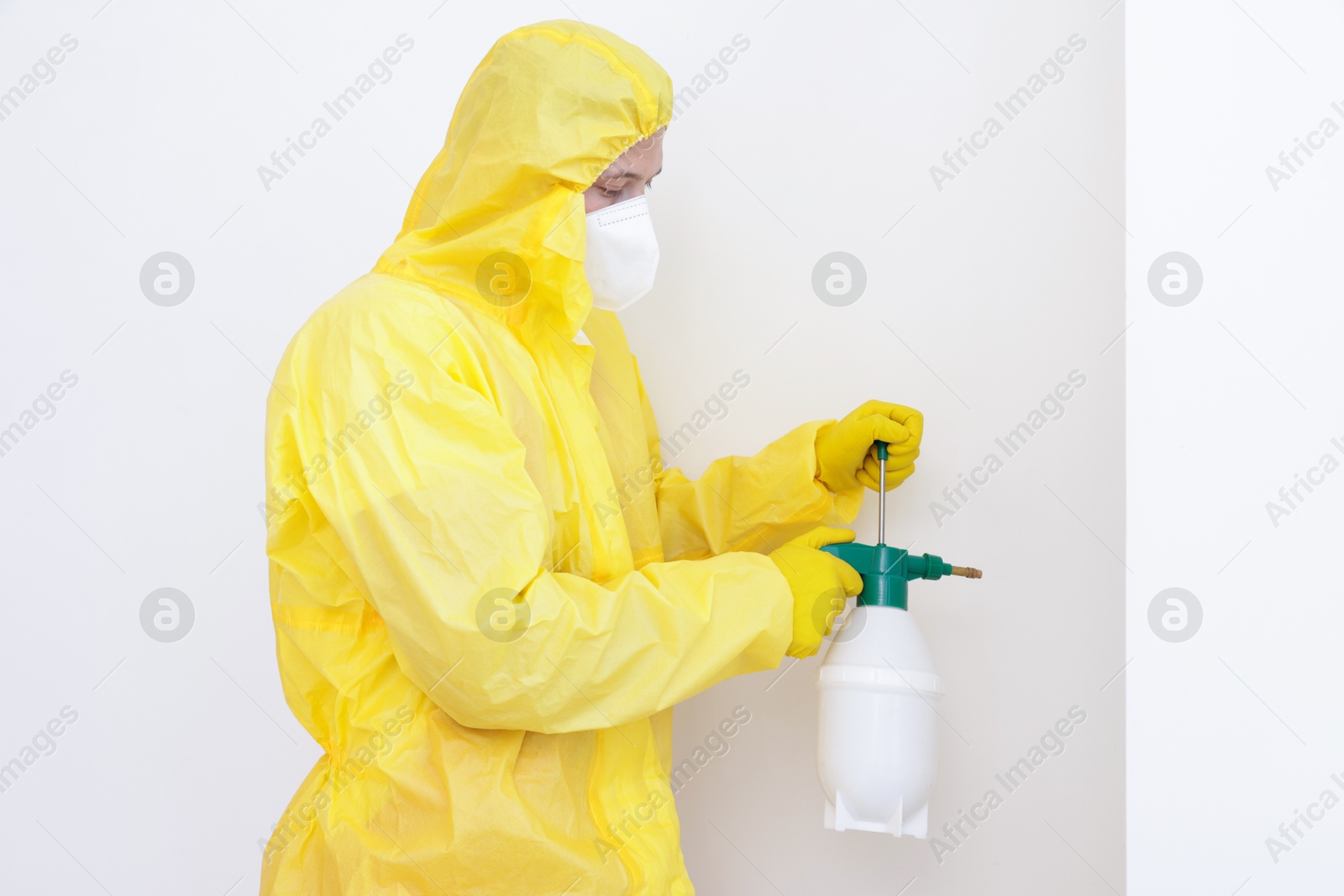 Photo of Pest control worker in protective suit spraying insecticide near white wall