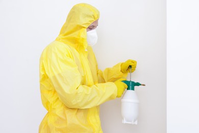 Photo of Pest control worker in protective suit spraying insecticide near white wall