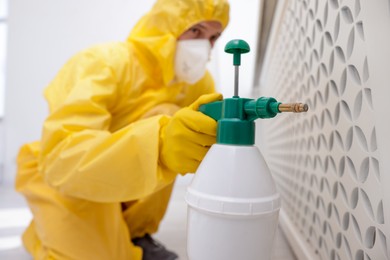 Photo of Pest control worker in protective suit spraying insecticide indoors, selective focus