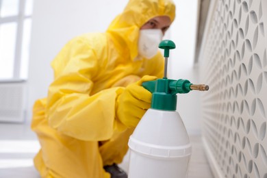 Photo of Pest control worker in protective suit spraying insecticide indoors, selective focus