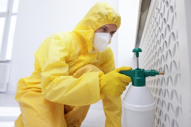 Photo of Pest control worker in protective suit spraying insecticide indoors, selective focus