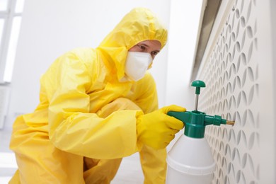 Photo of Pest control worker in protective suit spraying insecticide indoors, selective focus