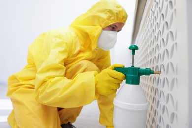 Photo of Pest control worker in protective suit spraying insecticide indoors, selective focus