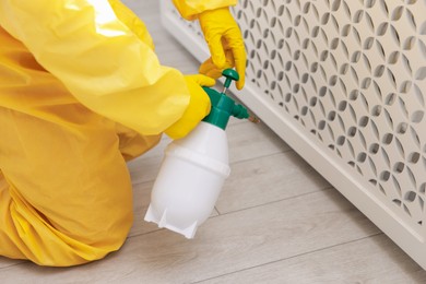 Photo of Pest control worker in protective suit spraying insecticide indoors, closeup