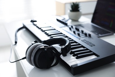 Photo of MIDI keyboard, headphones and laptop on white wooden table indoors, closeup