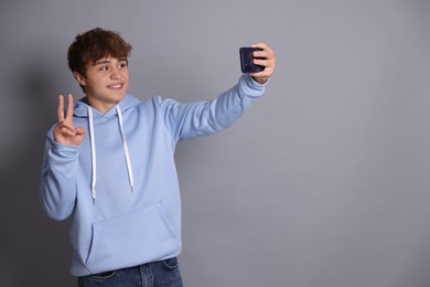 Photo of Smiling teenage boy taking selfie by smartphone and showing peace sign on grey background. Space for text