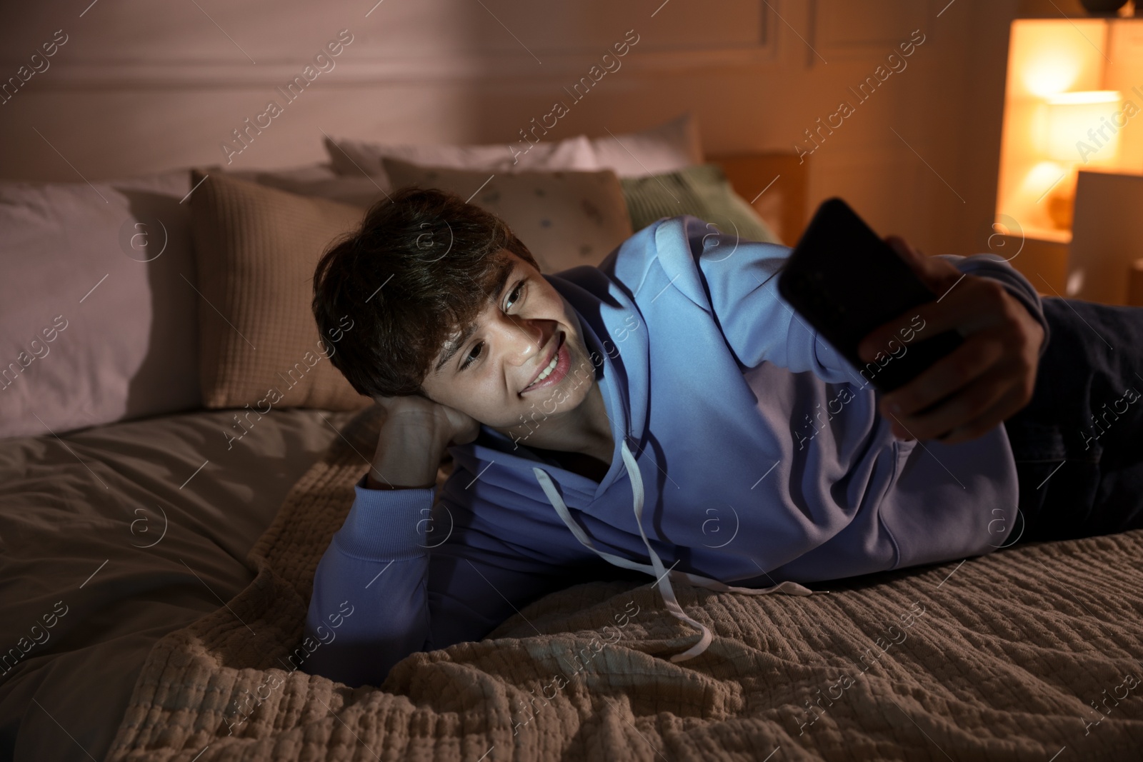 Photo of Smiling teenage boy having videocall by smartphone on bed at home