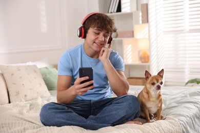 Photo of Smiling teenage boy in headphones using smartphone and dog on bed at home