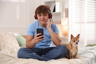 Photo of Smiling teenage boy in headphones using smartphone and dog on bed at home