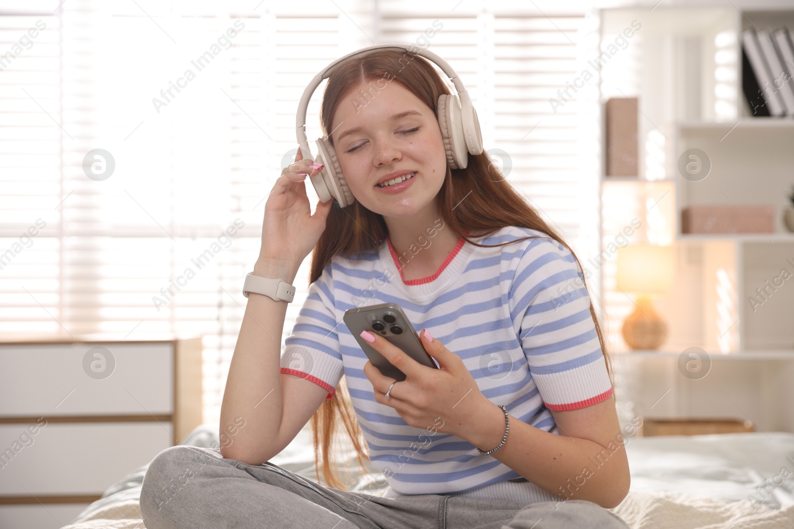 Smiling teenage girl in headphones with smartphone on bed at home Photo of Smiling teenage girl in headphones with smartphone on bed at home
