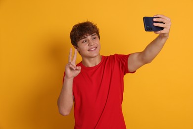 Photo of Smiling teenage boy taking selfie by smartphone and showing peace sign on yellow background