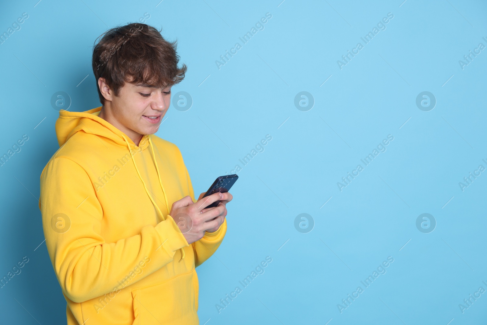 Photo of Smiling teenage boy using smartphone on light blue background. Space for text