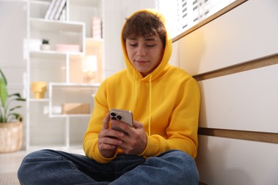 Photo of Teenage boy using smartphone near chest of drawers at home