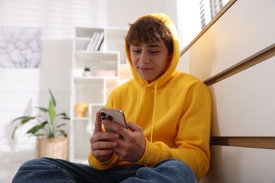 Photo of Teenage boy using smartphone near chest of drawers at home