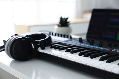 Photo of MIDI keyboard, headphones and laptop on white wooden table indoors, closeup
