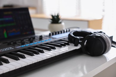 Photo of MIDI keyboard, headphones and laptop on white wooden table indoors, closeup