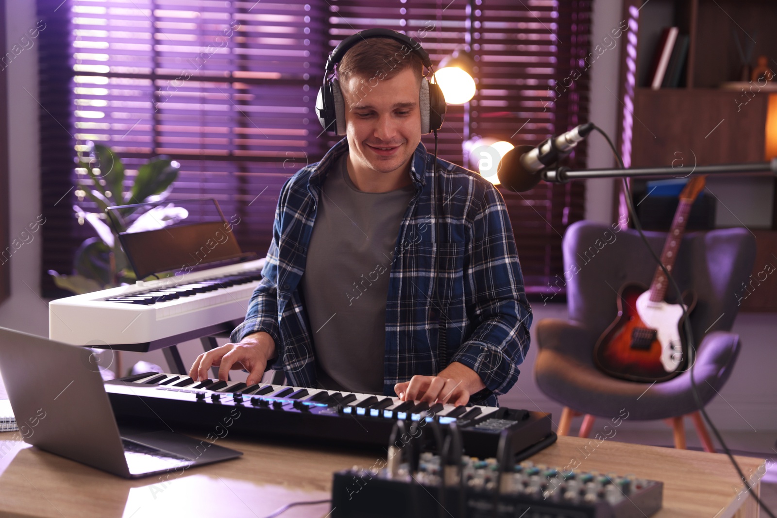 Photo of Smiling man in headphones working with MIDI keyboard at table in home studio