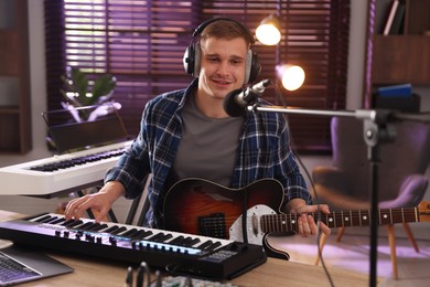 Photo of Smiling man in headphones working with MIDI keyboard and microphone at table in home studio