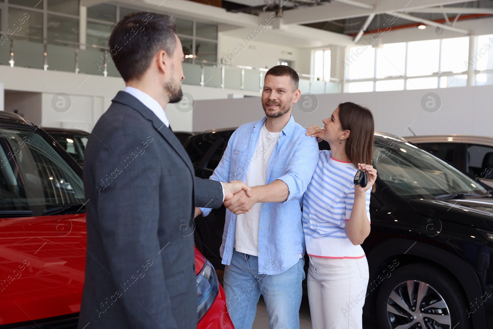 Photo of Salesman shaking hands with happy family near new car in salon
