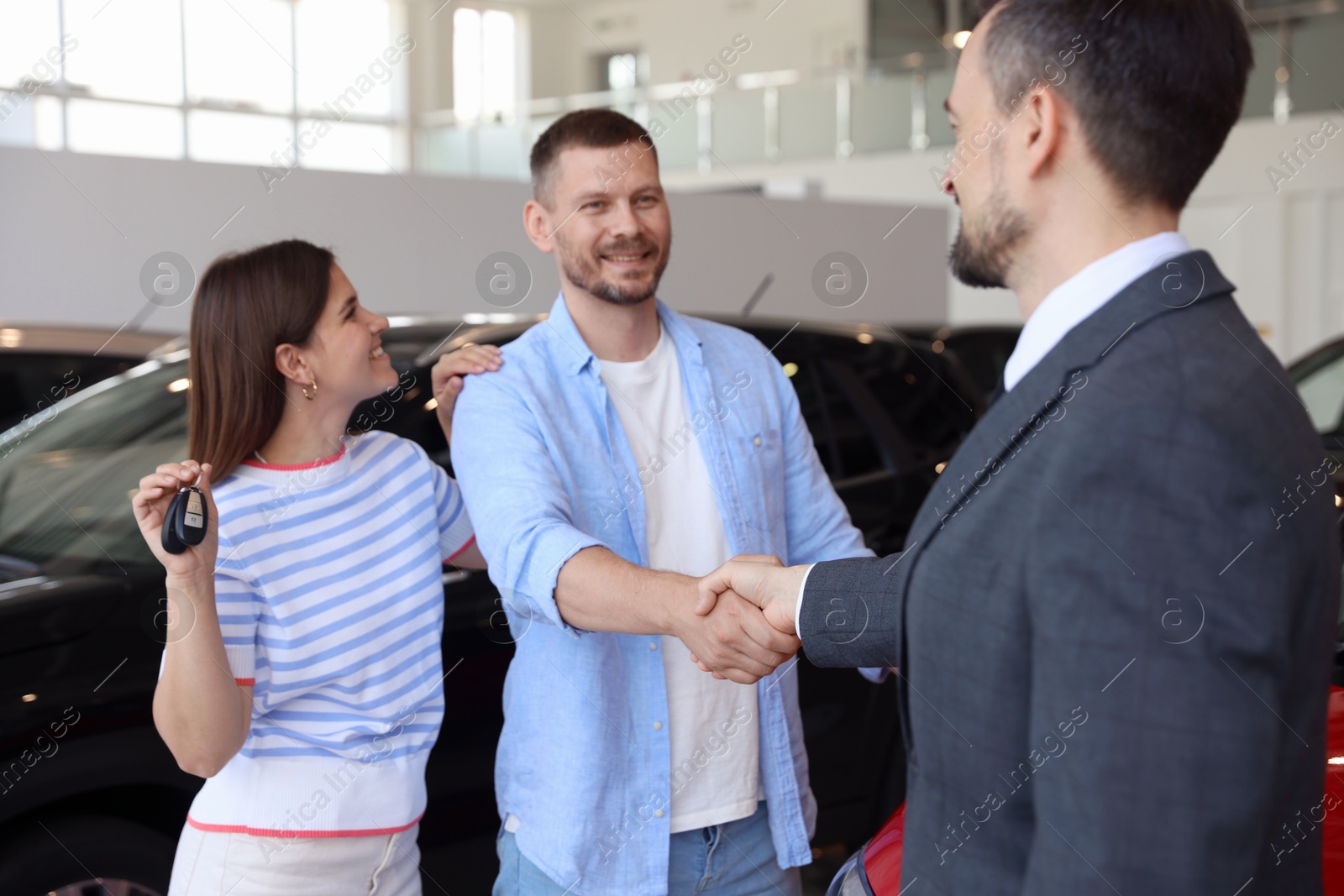 Photo of Salesman shaking hands with happy family near new car in salon