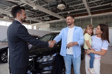 Photo of Salesman shaking hands with happy family near new car in salon