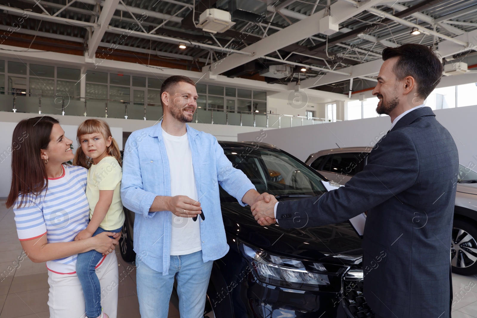 Photo of Salesman shaking hands with happy family near new car in salon