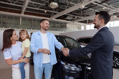 Photo of Salesman shaking hands with happy family near new car in salon