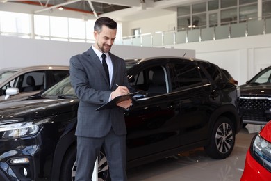 Photo of Smiling salesman working with clipboard near new car in salon