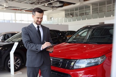Photo of Smiling salesman working with clipboard near new car in salon