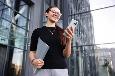 Photo of Successful businesswoman with smartphone and folder near office building, low angle view