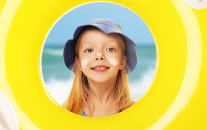 Image of Cute little girl with sunscreen on her face looking through hole of inflatable ring at beach. Sun protection while outdoor recreation in summer