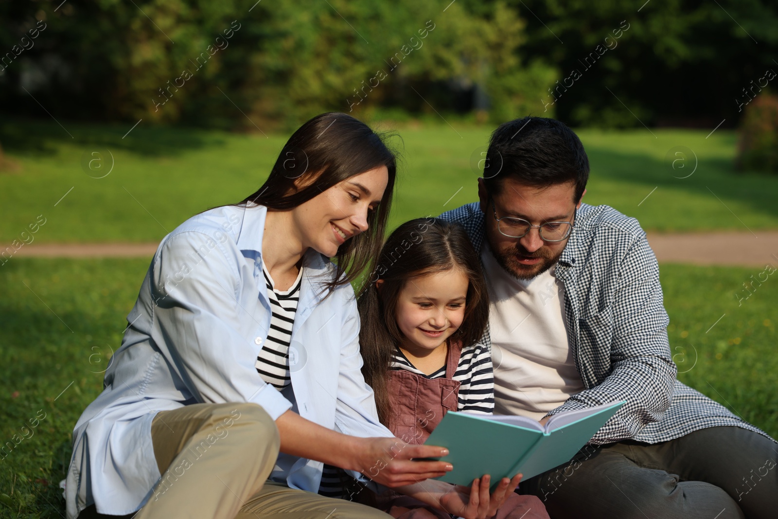 Photo of Family getaway. Happy parents and daughter reading book in park