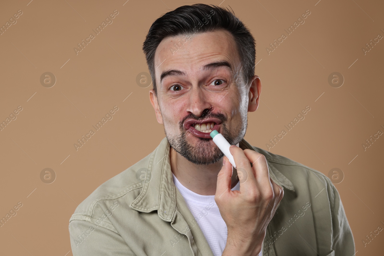 Funny man applying lip balm onto his lips on light brown background Photo of Funny man applying lip balm onto his lips on light brown background