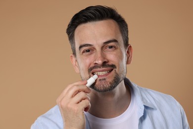 Photo of Smiling man applying lip balm onto his lips on light brown background