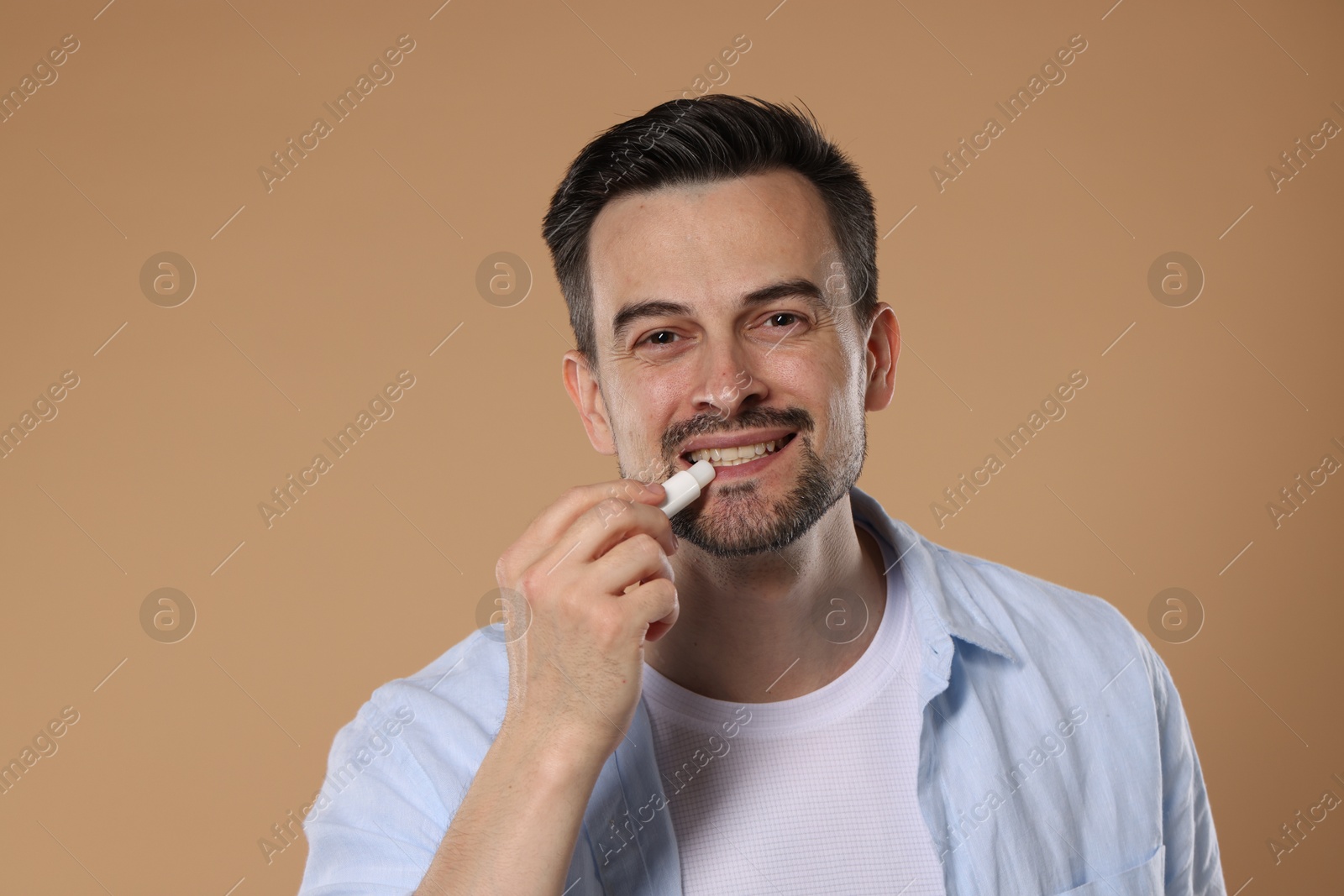 Smiling man applying lip balm onto his lips on light brown background Photo of Smiling man applying lip balm onto his lips on light brown background