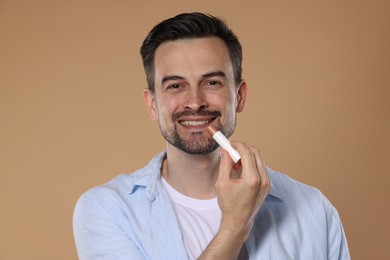 Photo of Smiling man applying lip balm onto his lips on light brown background