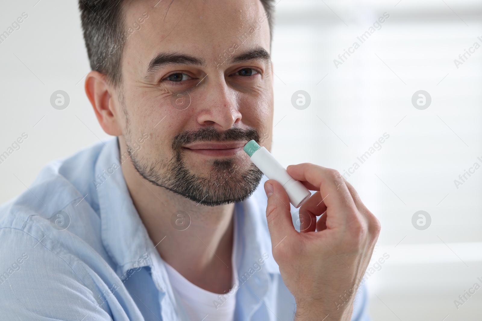 Photo of Handsome man applying lip balm onto his lips on blurred light background