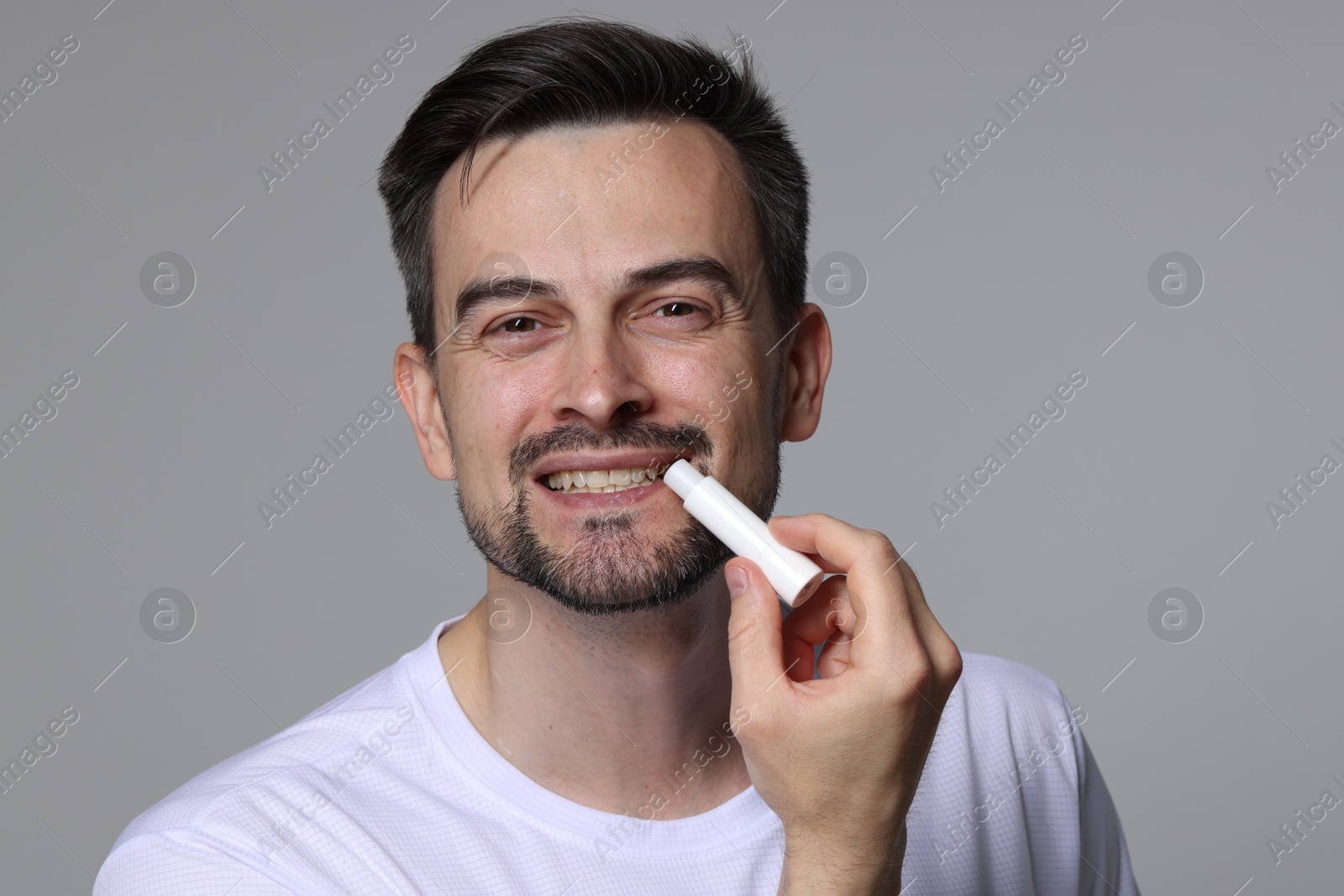 Smiling man applying lip balm onto his lips on grey background Photo of Smiling man applying lip balm onto his lips on grey background