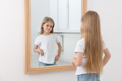 Photo of Upset little girl looking at herself in mirror indoors