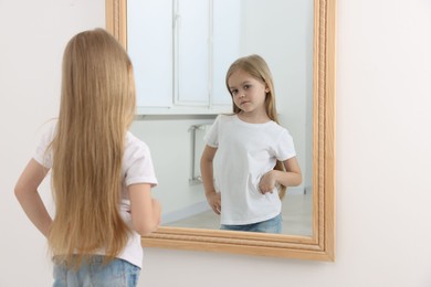 Photo of Upset little girl looking at herself in mirror indoors